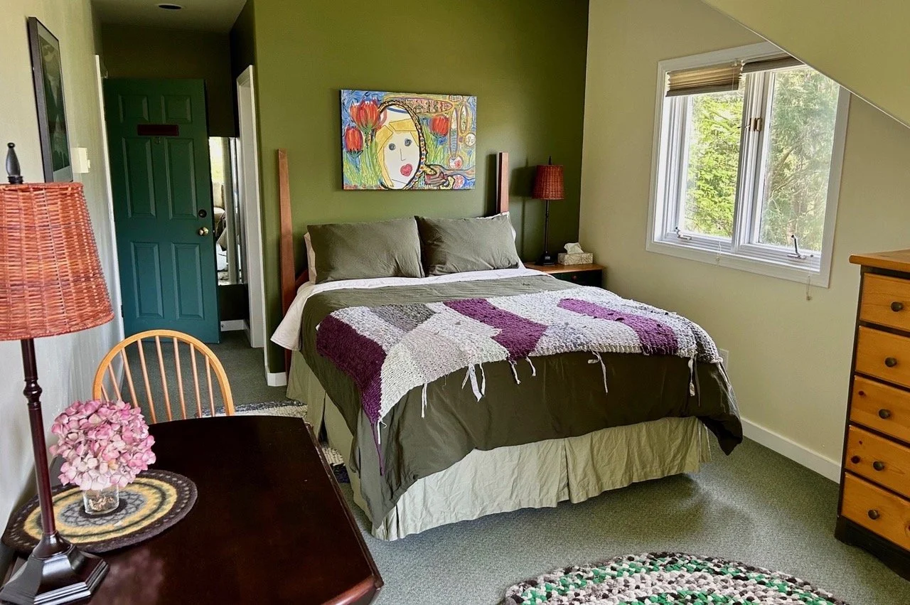 A cozy bedroom with a bed featuring brown pillows and a multicolored patchwork blanket. There is a window with a view of green trees, a wooden dresser, and a small table with a pink flower and a lampshade. A colorful abstract portrait hangs on the gr