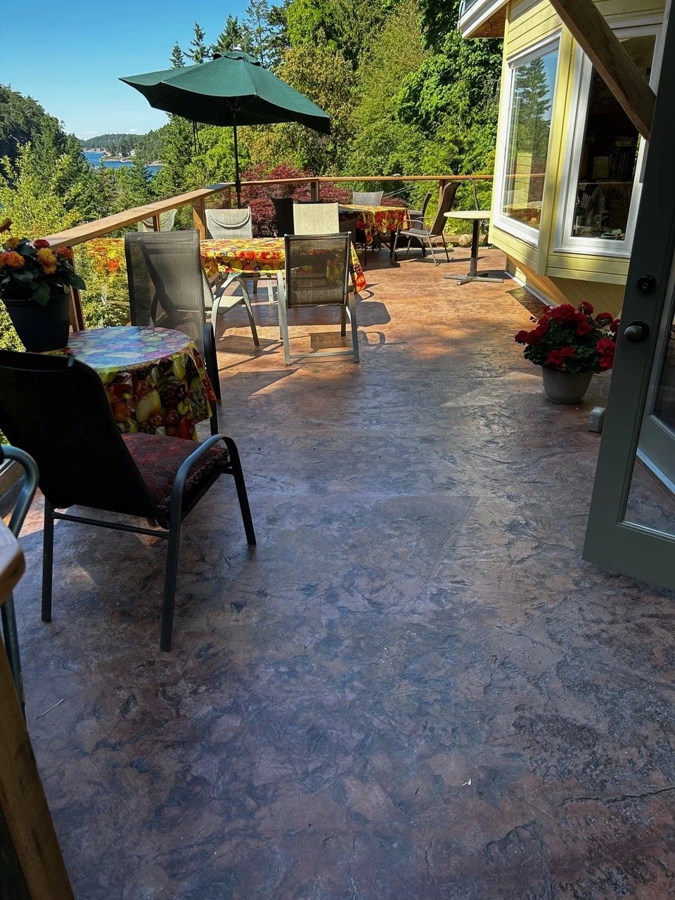 An outdoor patio with several tables and chairs, a large green umbrella, potted flowers, and a view of trees and water in the distance.