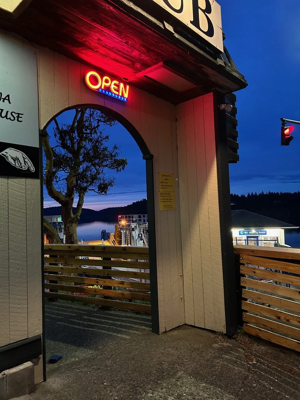 View of a waterfront dock area through an arched doorway with a red 'Open' sign hanging above, during dusk with a tree and hills in the background.