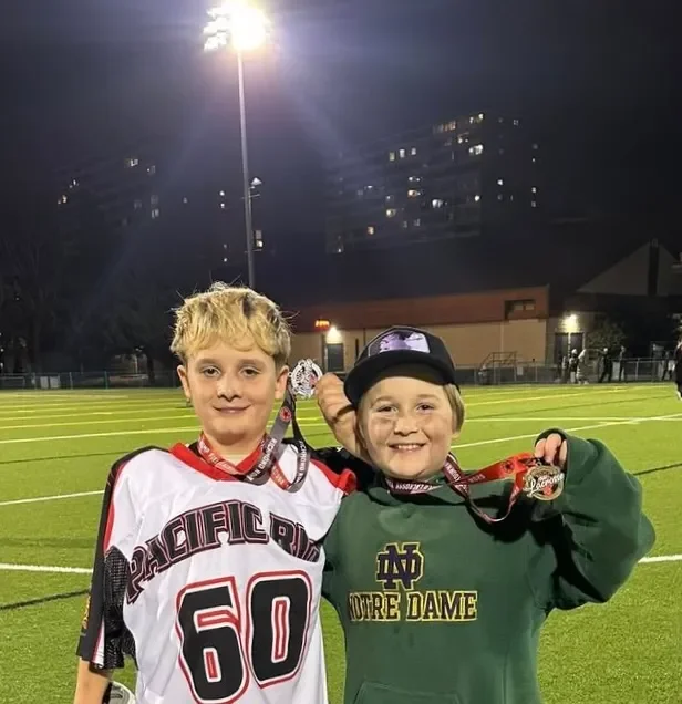 Two young boys standing on a lit football field at night, smiling and displaying medals around their necks. One wears a white football jersey with the number 60, and the other wears a green Notre Dame sweatshirt and a black cap.