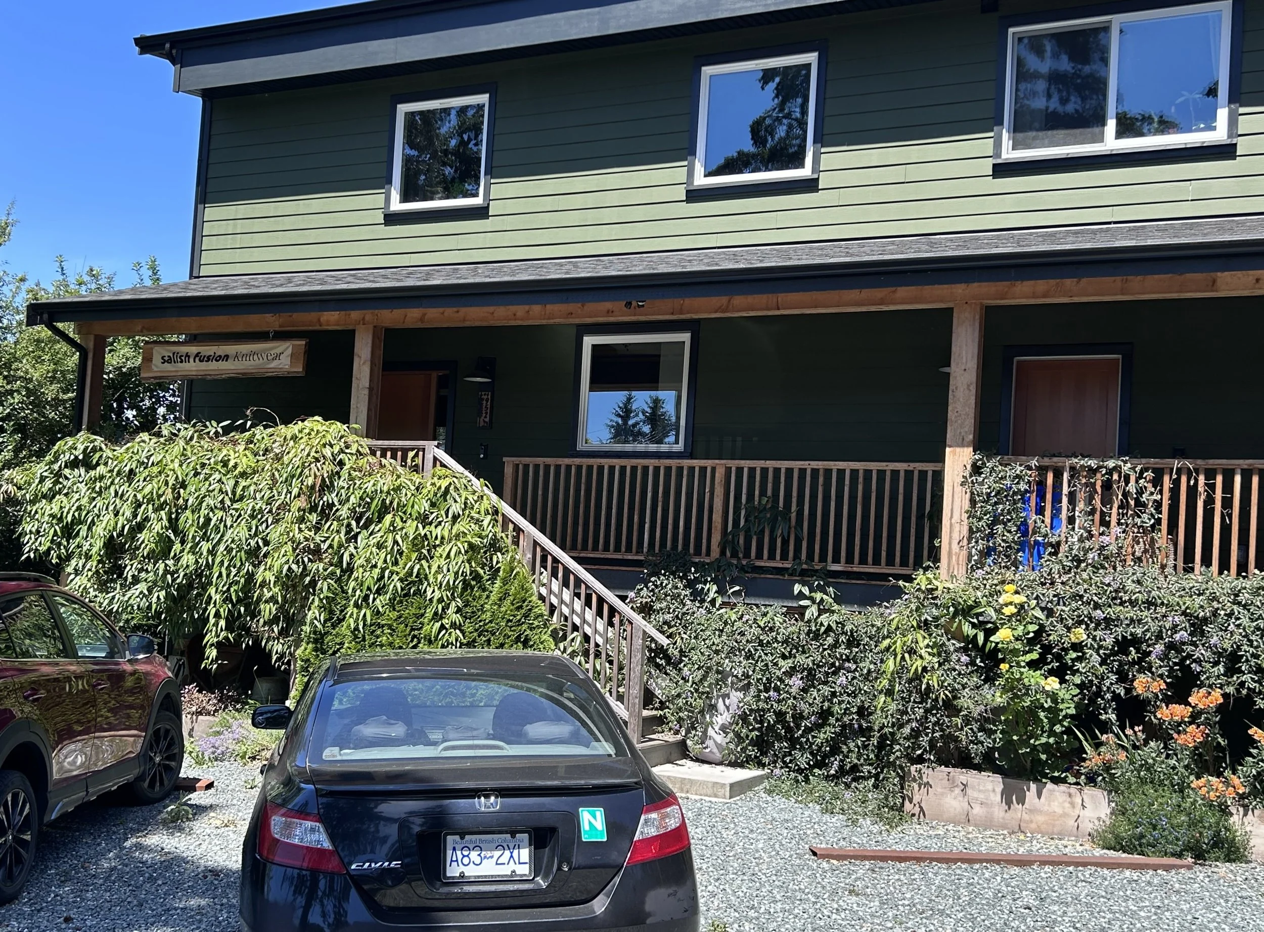 A two-story house painted in green with white window frames and black trim. The house has a wooden porch with a sign that reads 'Salish Fusion Knitwear.' In front of the house, there are several cars parked, including a black car with a license plate from British Columbia and an 'N' sticker. The yard is landscaped with bushes, flowers, and trees.