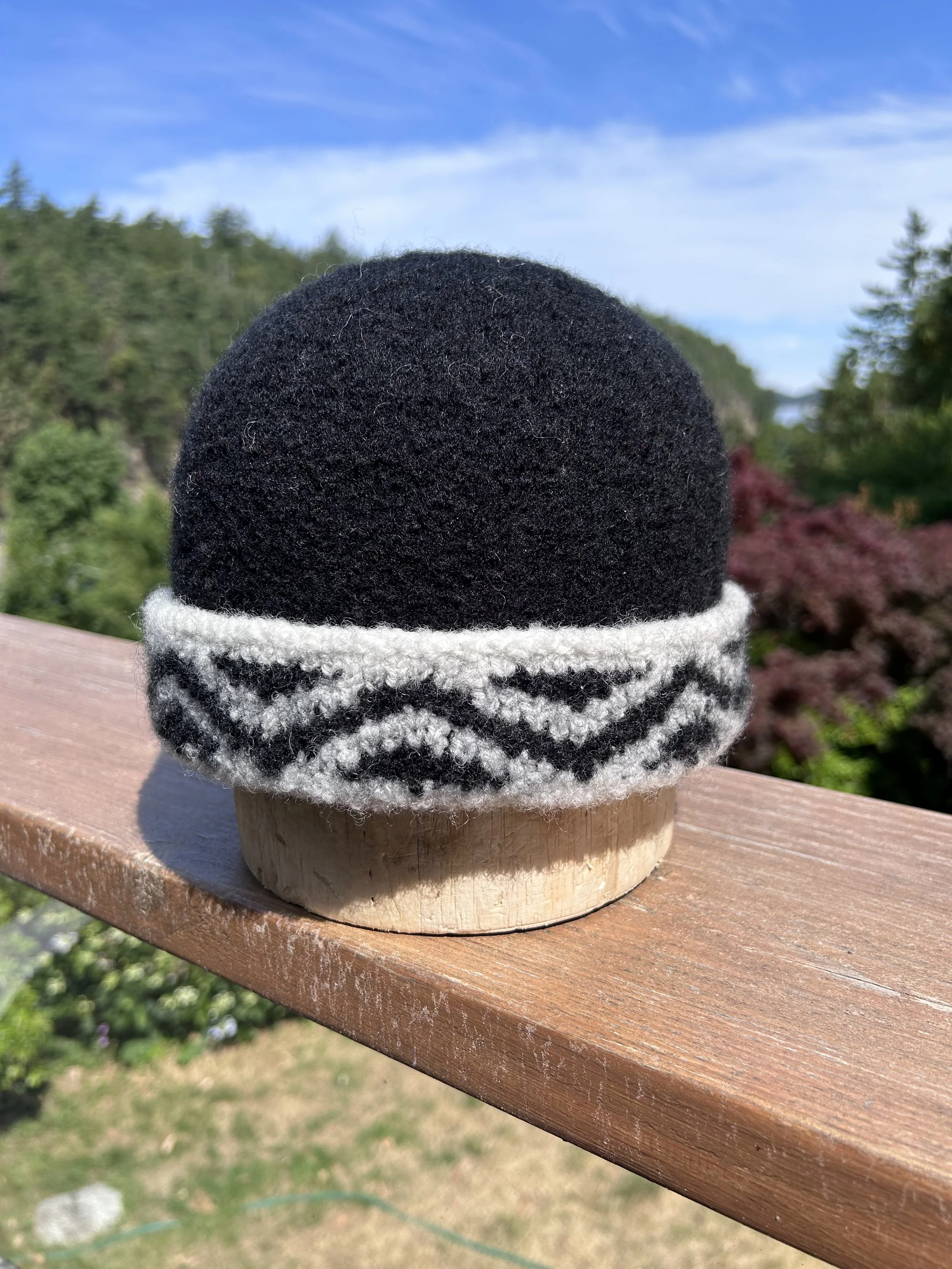 Close-up of a black knit hat with a white and black geometric pattern, placed on a wooden surface outdoors with trees and blue sky in the background.