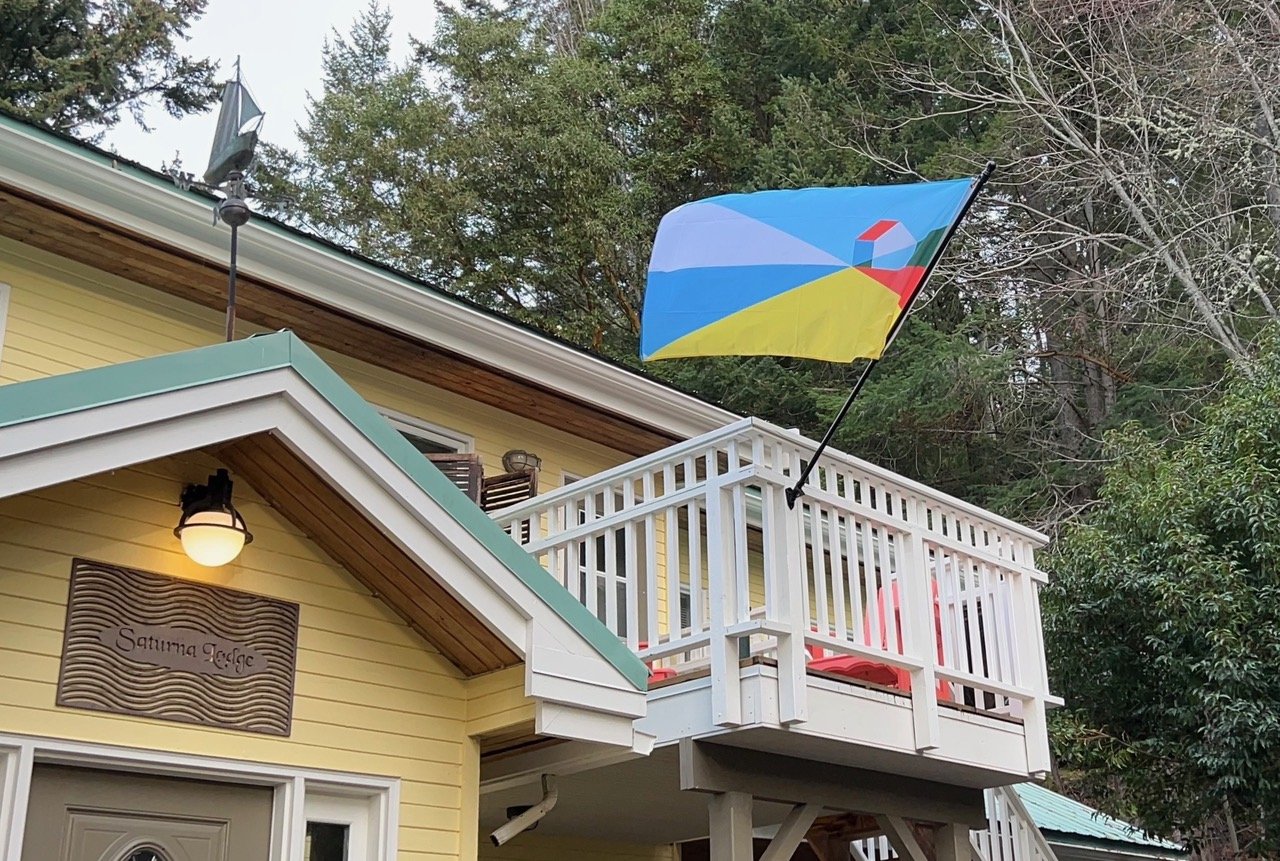 Yellow house with a sign that says 'Saturna Lodge', a balcony with red chairs, and a colorful geometric-patterned flag attached to the balcony above the sign.