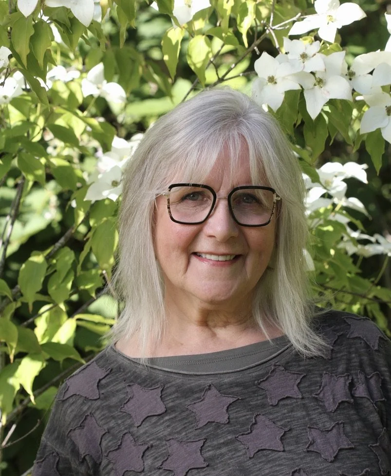 A smiling woman with glasses standing outdoors in front of white flowering plant, wearing a dark gray top with star patterns.