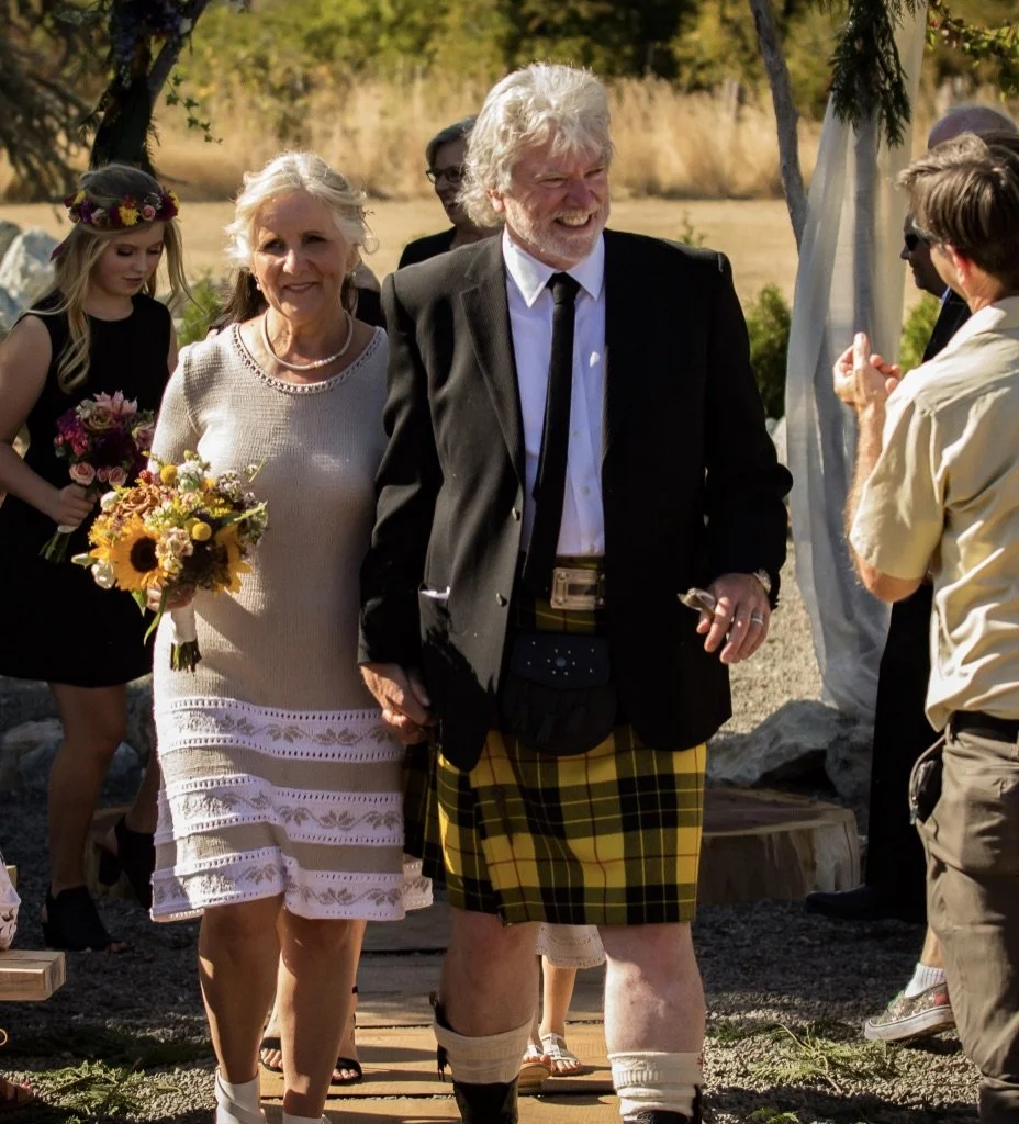 A man and a woman walking together at an outdoor event, with the woman holding a bouquet of flowers. The man is wearing a dark jacket, white shirt, and a yellow plaid kilt. The woman is wearing a white dress and a pearl necklace. There are other people in the background, some clapping and one woman wearing a flower crown, with trees and greenery around.