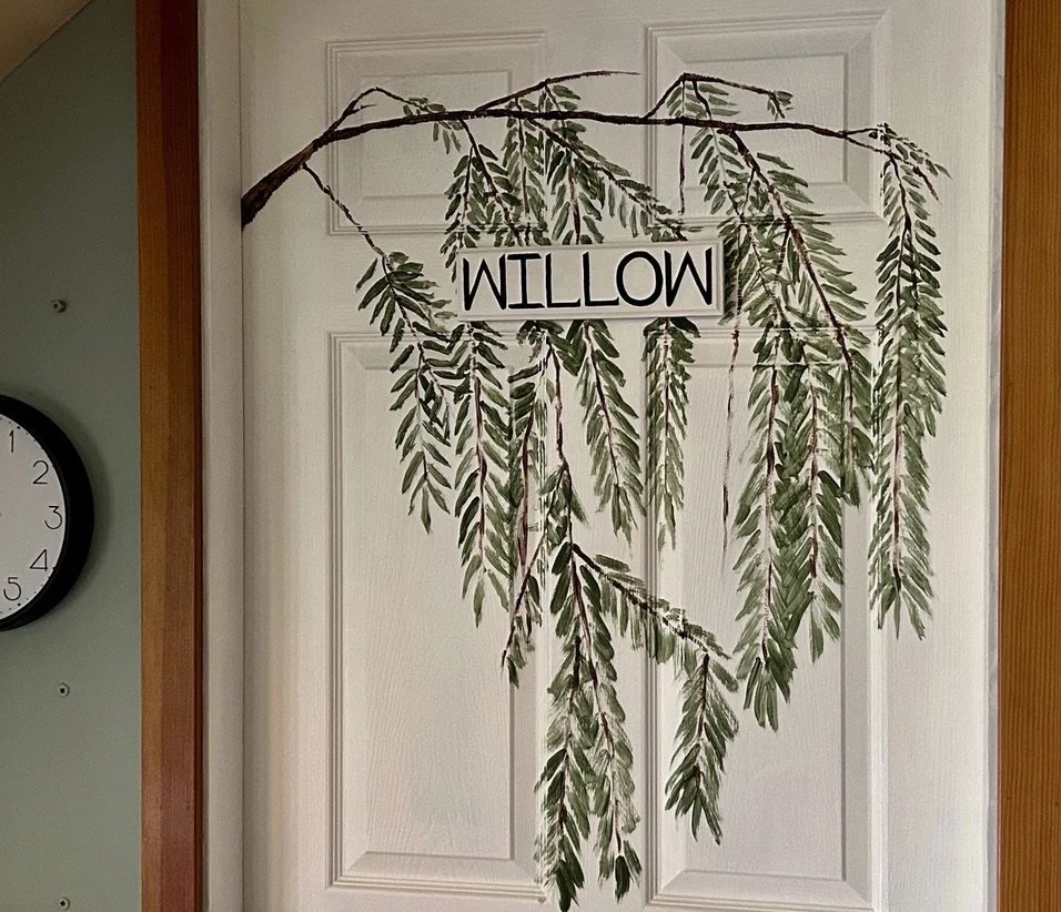 Decorative wall hanging with green leafy branches and a sign that says 'WILLOW' in black letters.