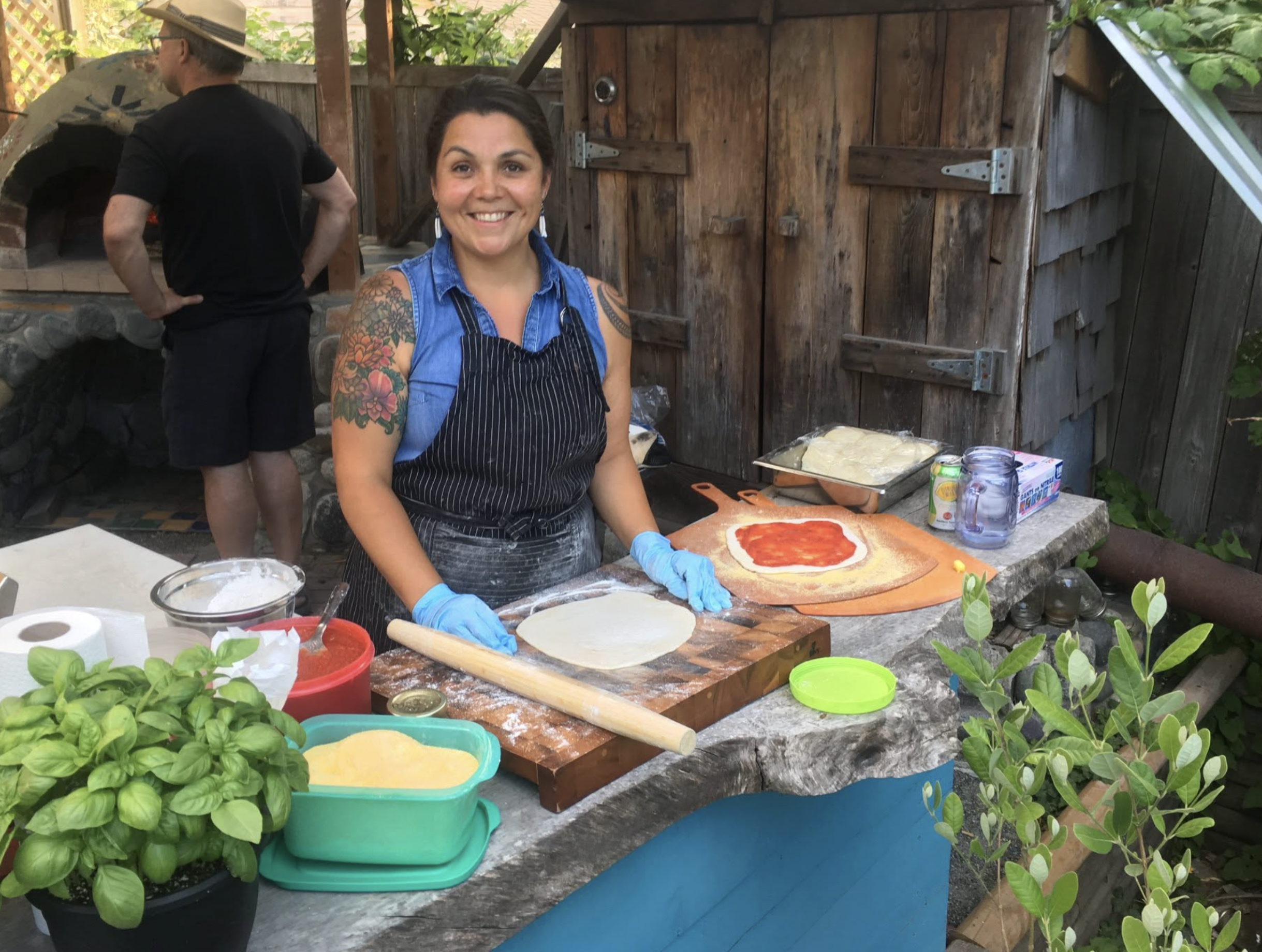 A woman with tattoos smiling while preparing pizza dough outdoors, with basil plants and cooking ingredients nearby, and a man in the background cooking at an outdoor brick oven.