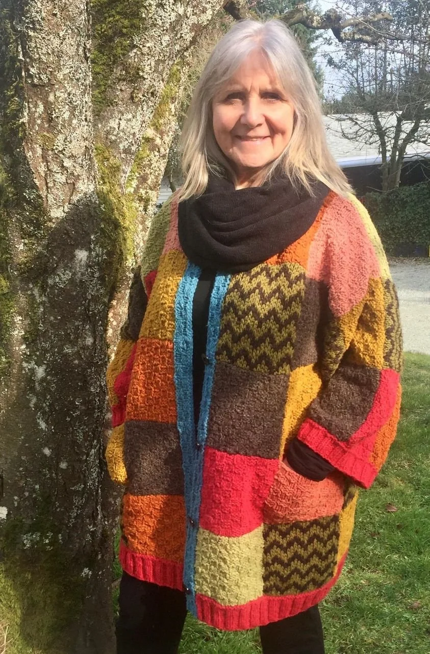 A smiling woman with gray hair wearing a colorful patchwork sweater and a black scarf, standing outdoors next to a tree with moss and lichen.