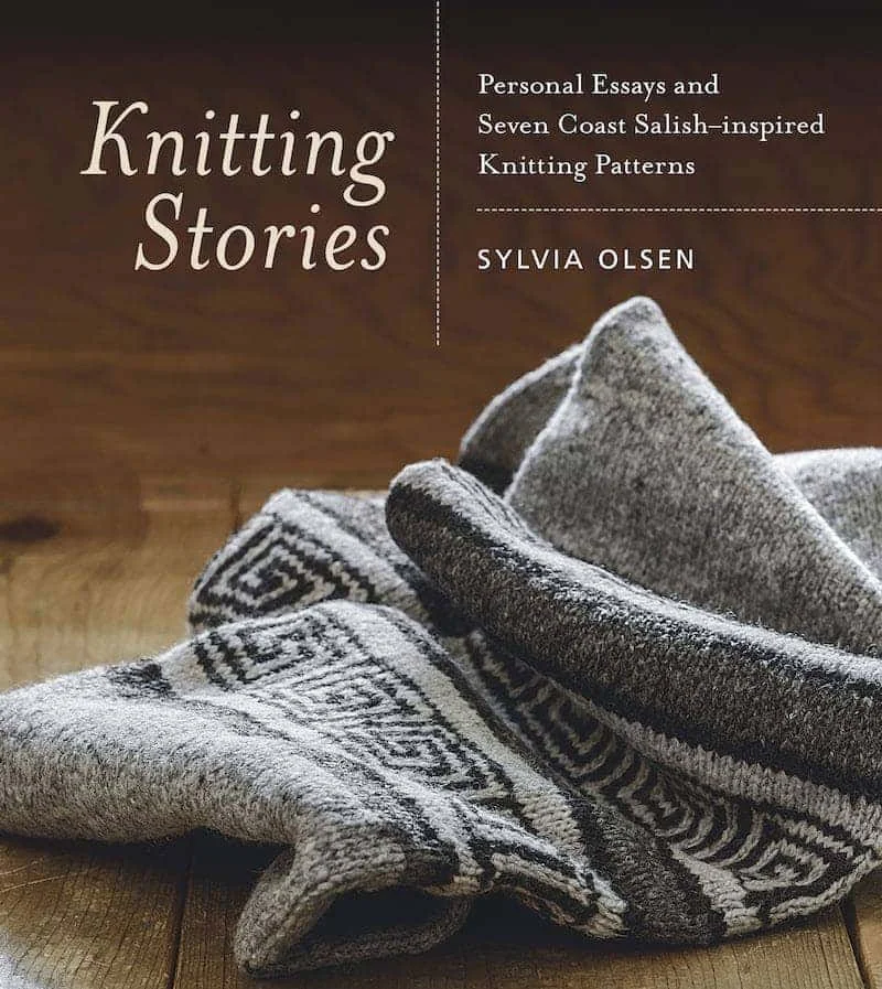 Cover of a book titled 'Knitting Stories' with a woolen blanket in gray and black colors folded on a wooden surface.