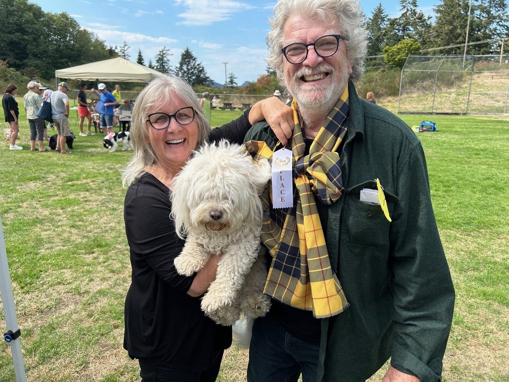 Smiling woman holding a fluffy white dog, standing beside a smiling man with glasses and a plaid scarf, at an outdoor event on a grassy field with other people and dogs in the background.
