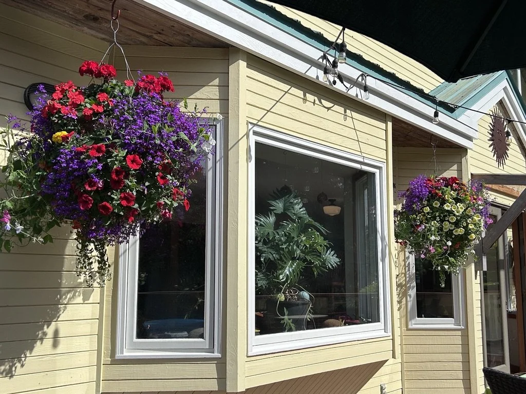Two hanging flower baskets with pink, purple, red, and white flowers on a yellow house exterior with large windows.