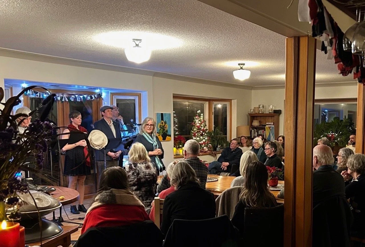 People gathered in a warmly decorated living room for a Christmas gathering, with some performing music on a small stage, and a Christmas tree in the background.