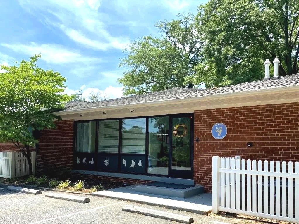 A brick building with large glass windows decorated with holiday-themed window stickers, a wreath on the door, and a sign reading 'galeopod Montessori.' The building is surrounded by trees and has a white picket fence.