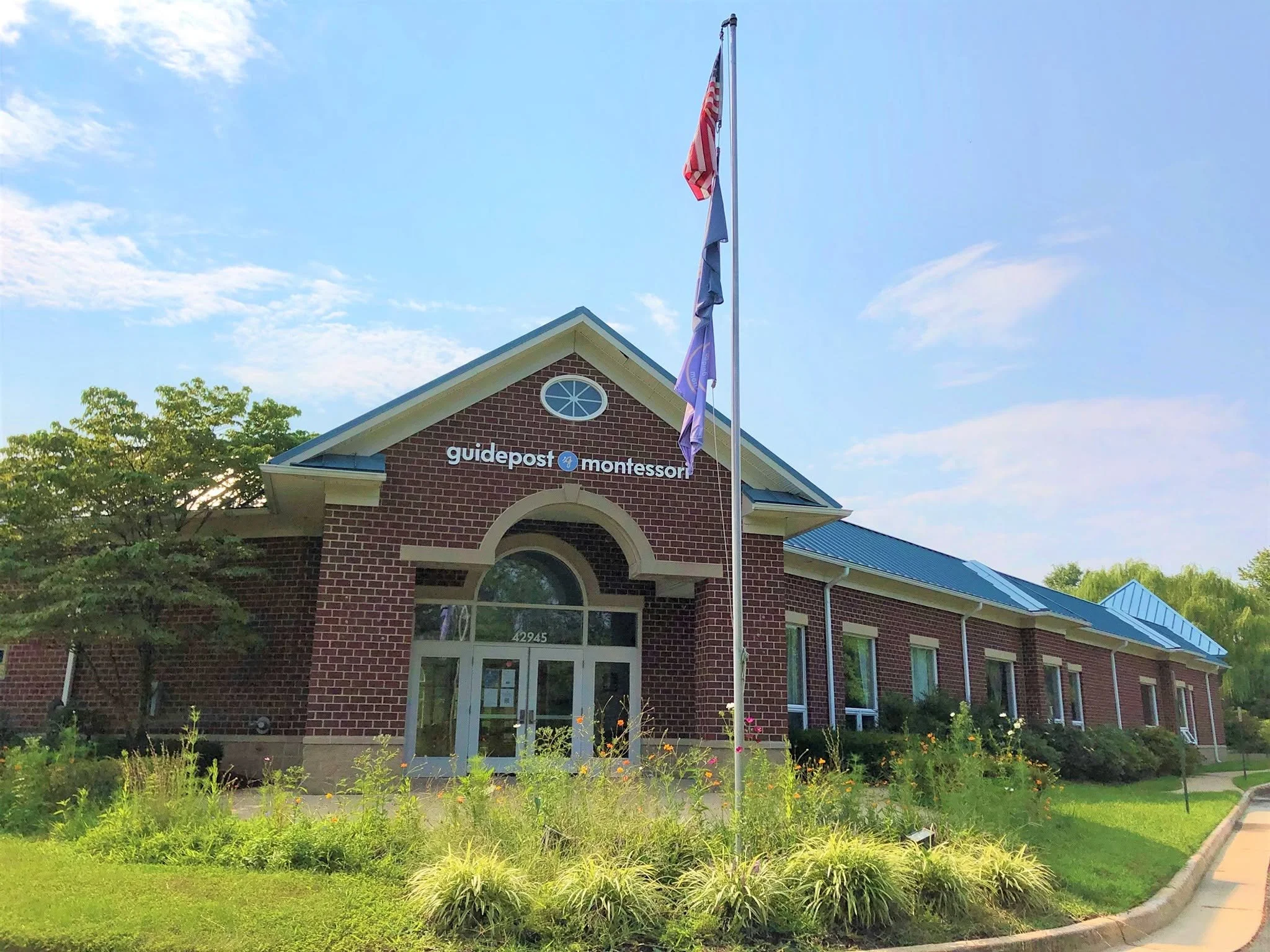Front view of Guidepost Montessori building with American flag and flags, surrounded by landscaped garden and trees, under a blue sky with clouds.