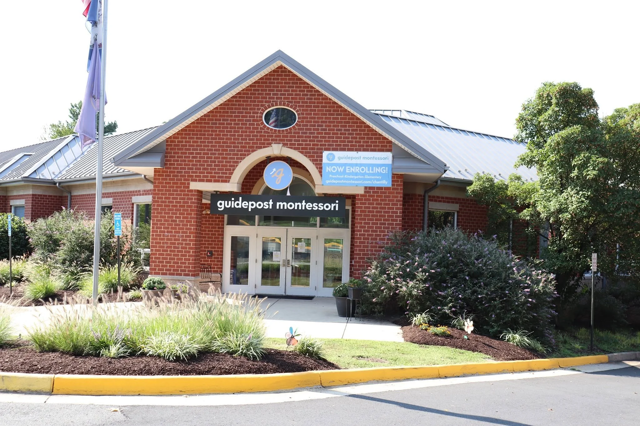 Red brick building with a gray metal roof and a sign that reads 'guidepost montessori' and 'NOW ENROLLING!' in front of a landscaped area with purple flowering bushes and a flagpole with flags.