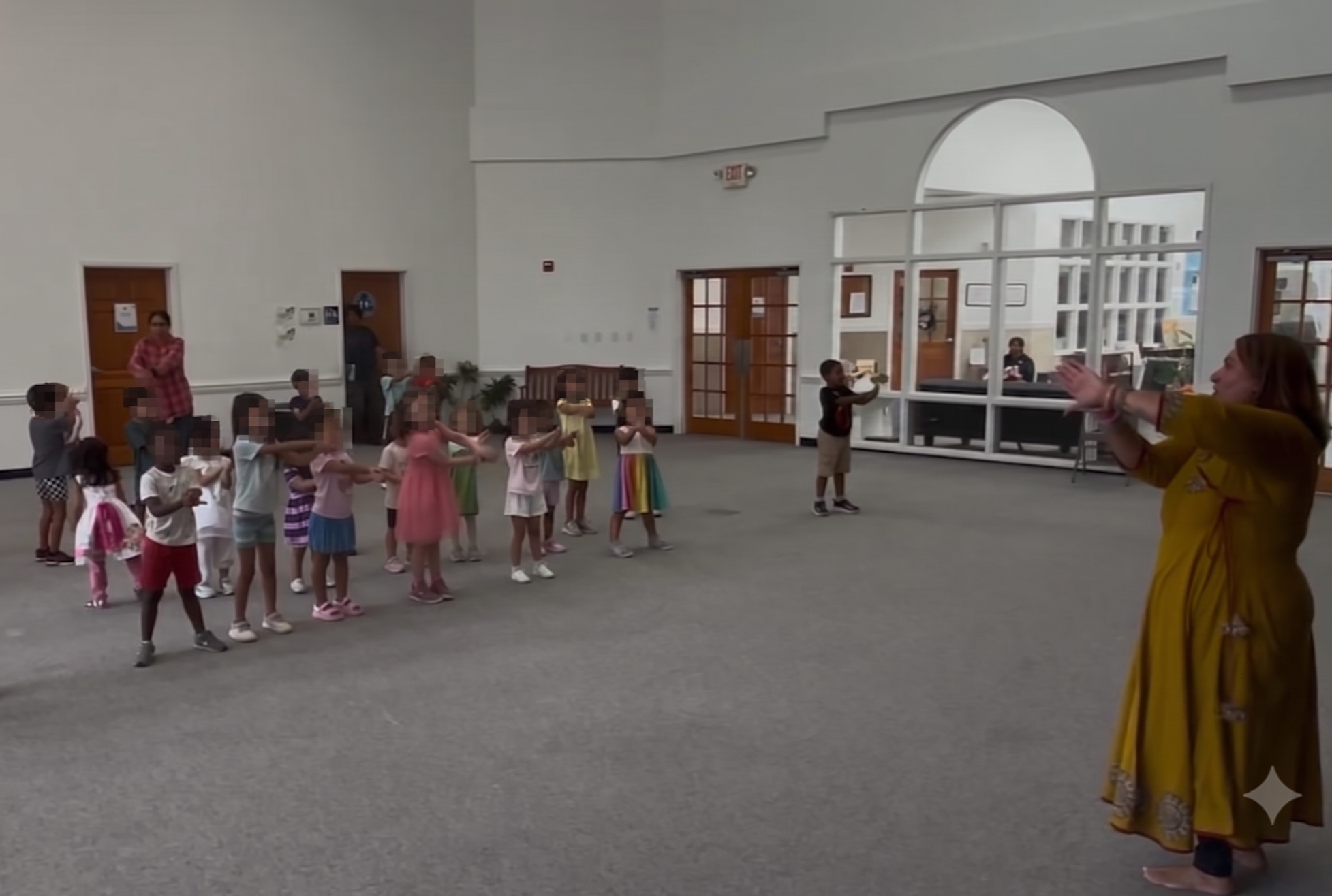 A group of children and an adult woman participating in an indoor activity in a spacious room with gray carpet and white walls, with some windows and framed pictures in the background.