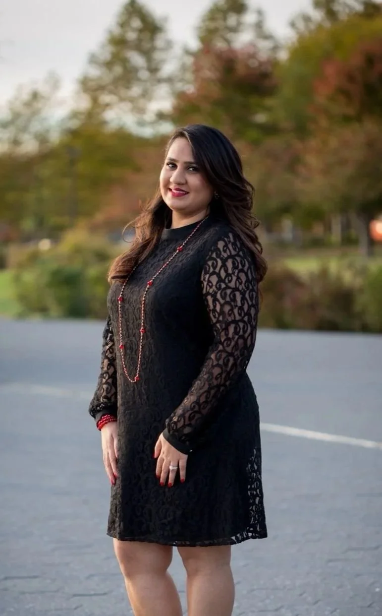 A woman in a black lace dress standing outdoors on a road with trees and fall foliage in the background.