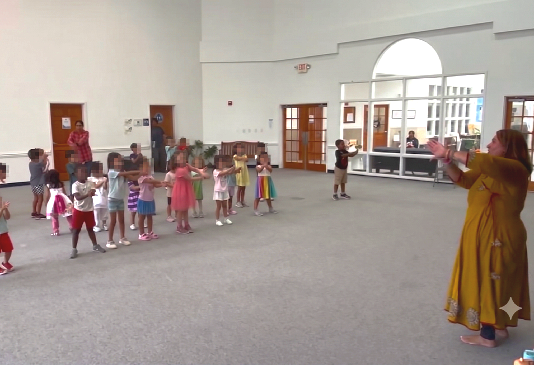 Children and a woman participating in a group activity, standing in a spacious room with large windows and white walls, possibly in a school or community center.