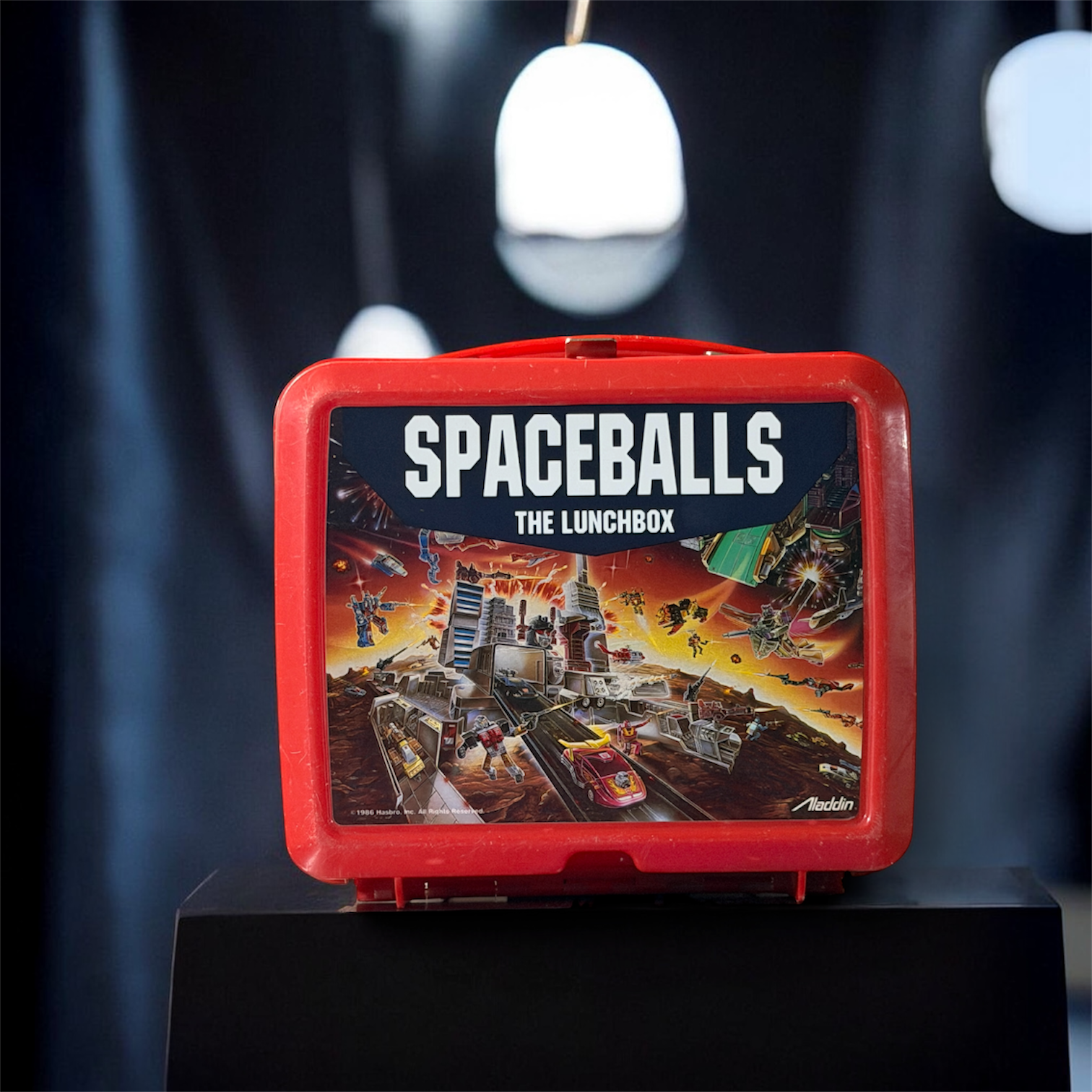 A red lunchbox with a 'Spaceballs' themed graphic on the front, featuring a space battle scene with various vehicles and characters from the movie, labeled 'The Lunchbox'.