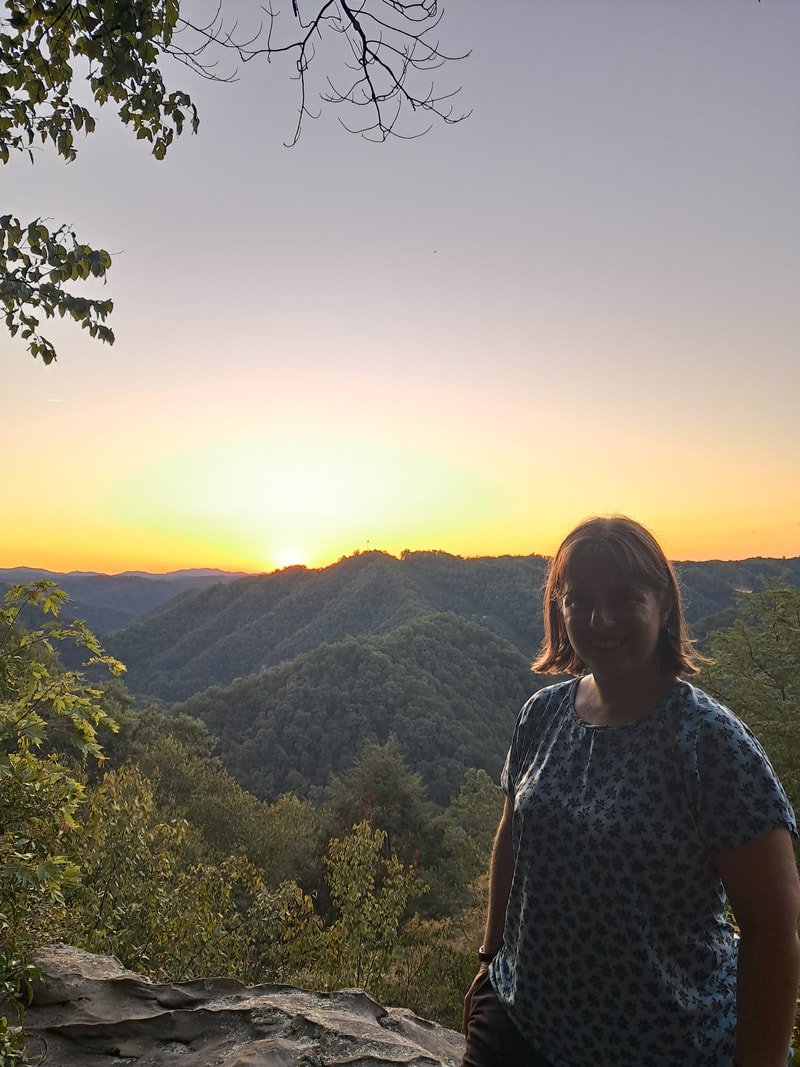 White woman stands in front of a mountain range while the sun sets behind them