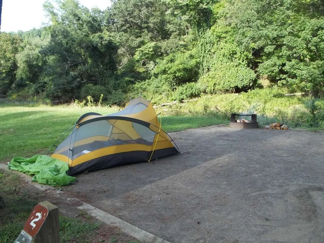 A small yellow and grey tent on a gravel camping pad overlooks a grassy bank and green trees