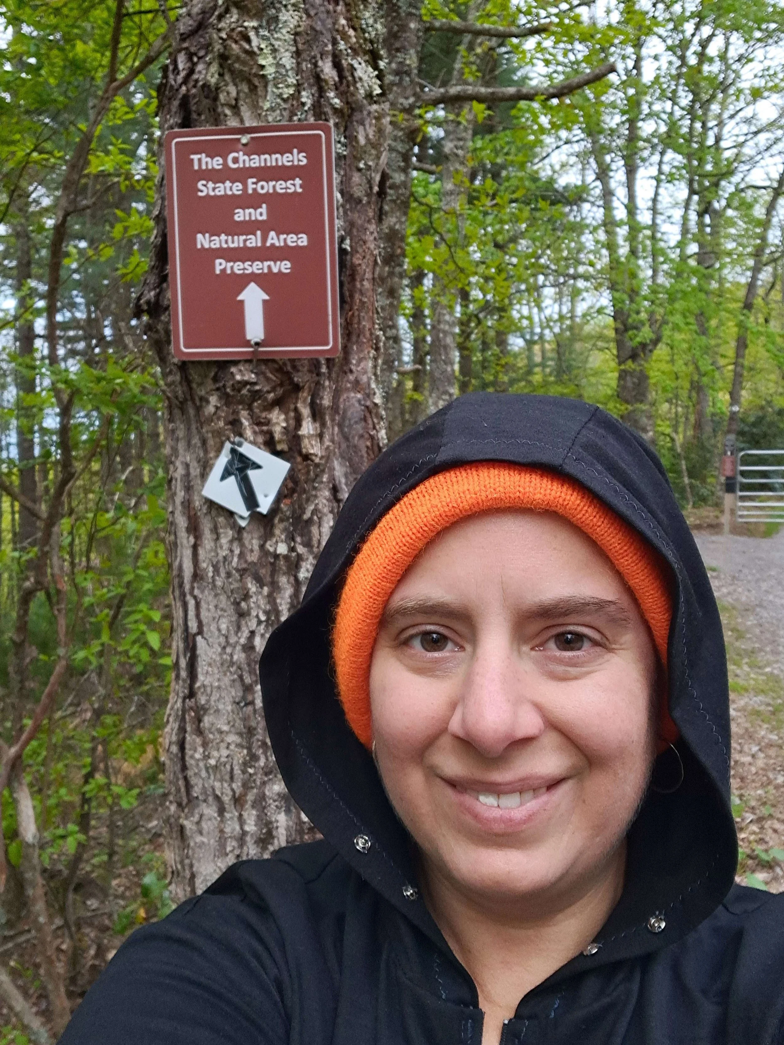 A white woman in an orange beanie and black hoodie stands in front of a sign for The Channels State Forest