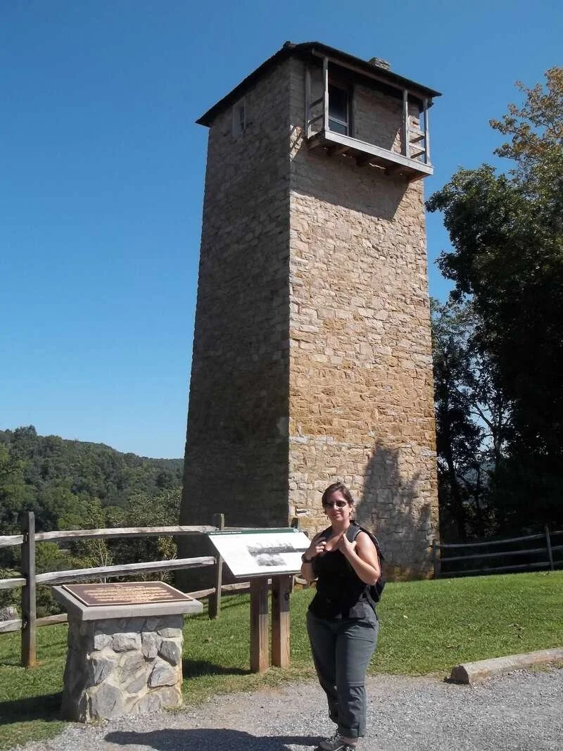 A short white woman in hiking clothes stands in front of a square stone tower