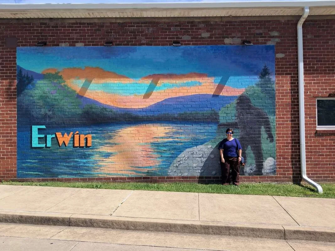 A white woman stands at a mural of a lake, Sasquatch, and the word Erwin