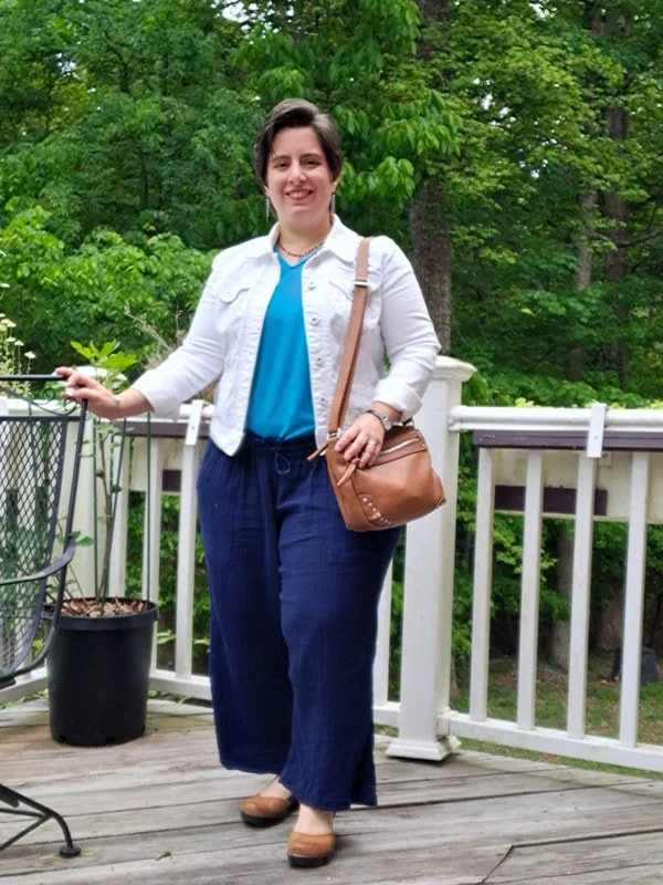 Woman wearing white jacket, bright blue t-shirt, navy drawstring pants, tan shoes, and a tan bag