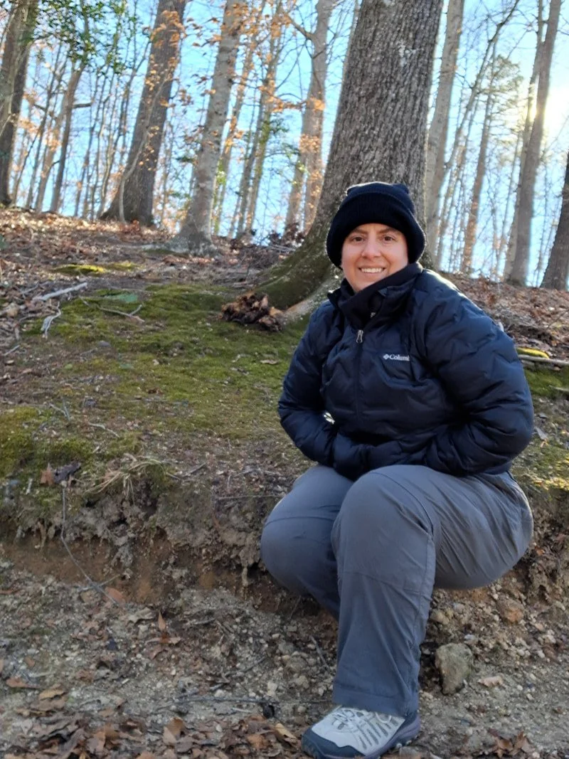 A white woman wearing a black winter hat and coat crouches in front of a mossy bank in a winter decidious forest