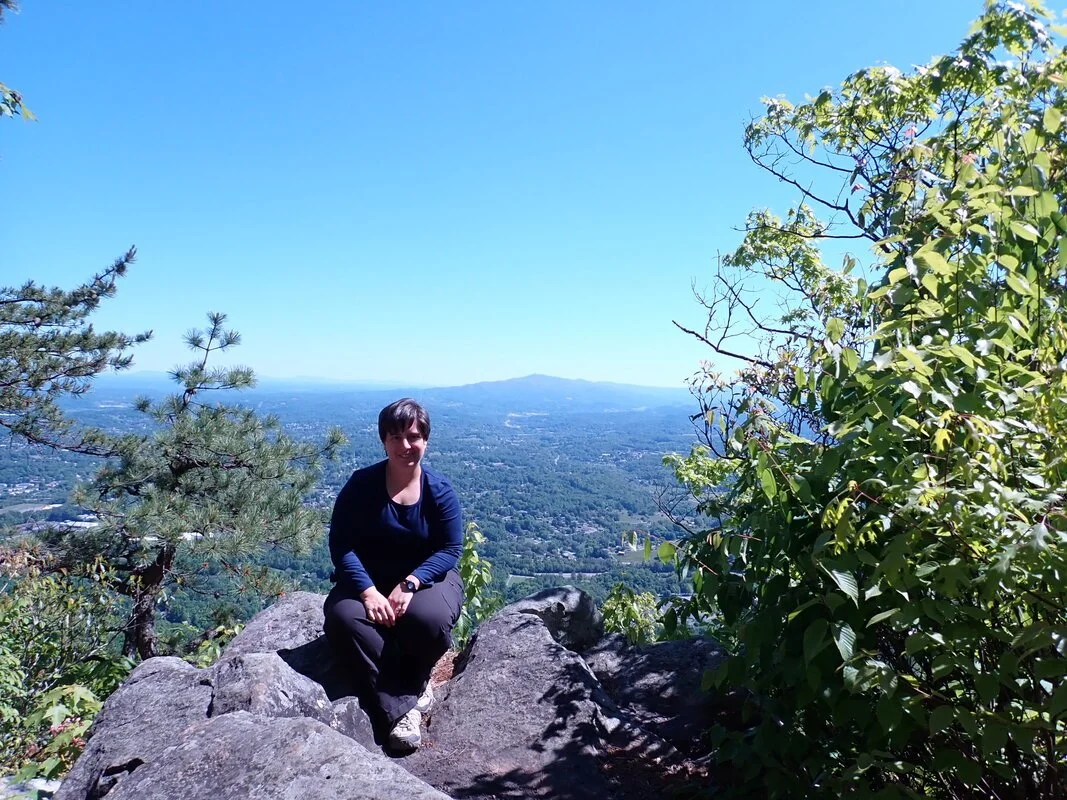 A white woman sits facing the camera, surrounded by trees and with a mountain peak in the distance behind her