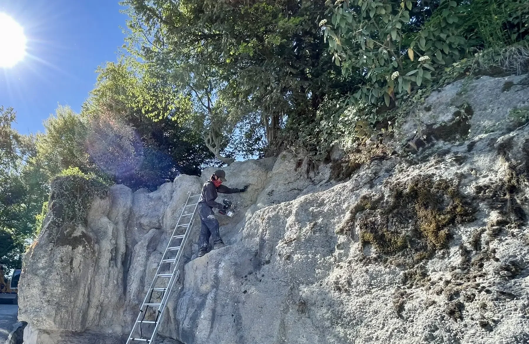 Ein Mitarbeiter koloriert die rekonstruierte Felswand am Gubelstein. Er steht auf einer Leiter zwischen den hellen Felsformationen; Teile des Felsens sind bereits von Moos und Vegetation bewachsen.