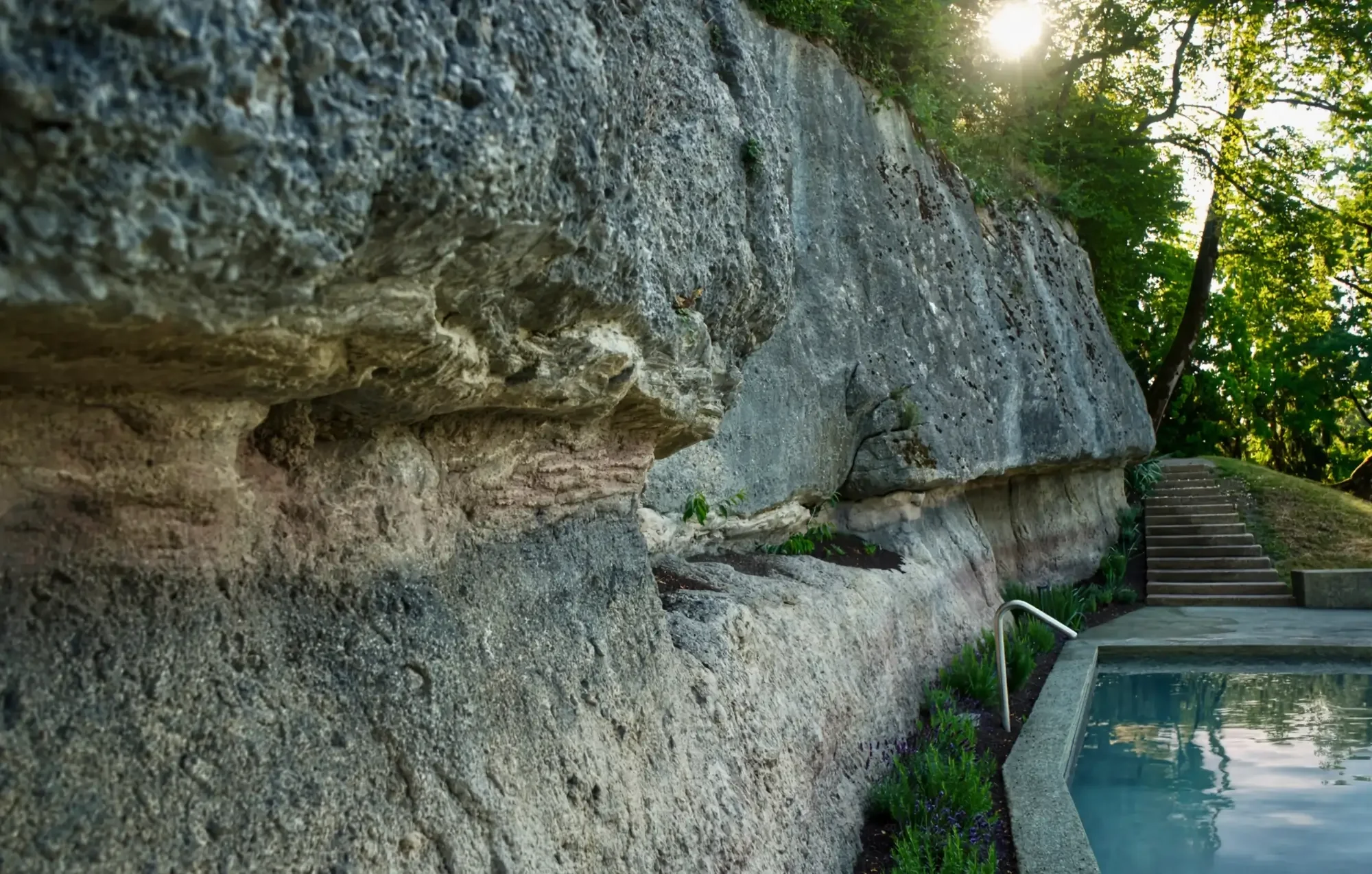 Felswand entlang eines Pools mit Treppe und Bäumen im Hintergrund