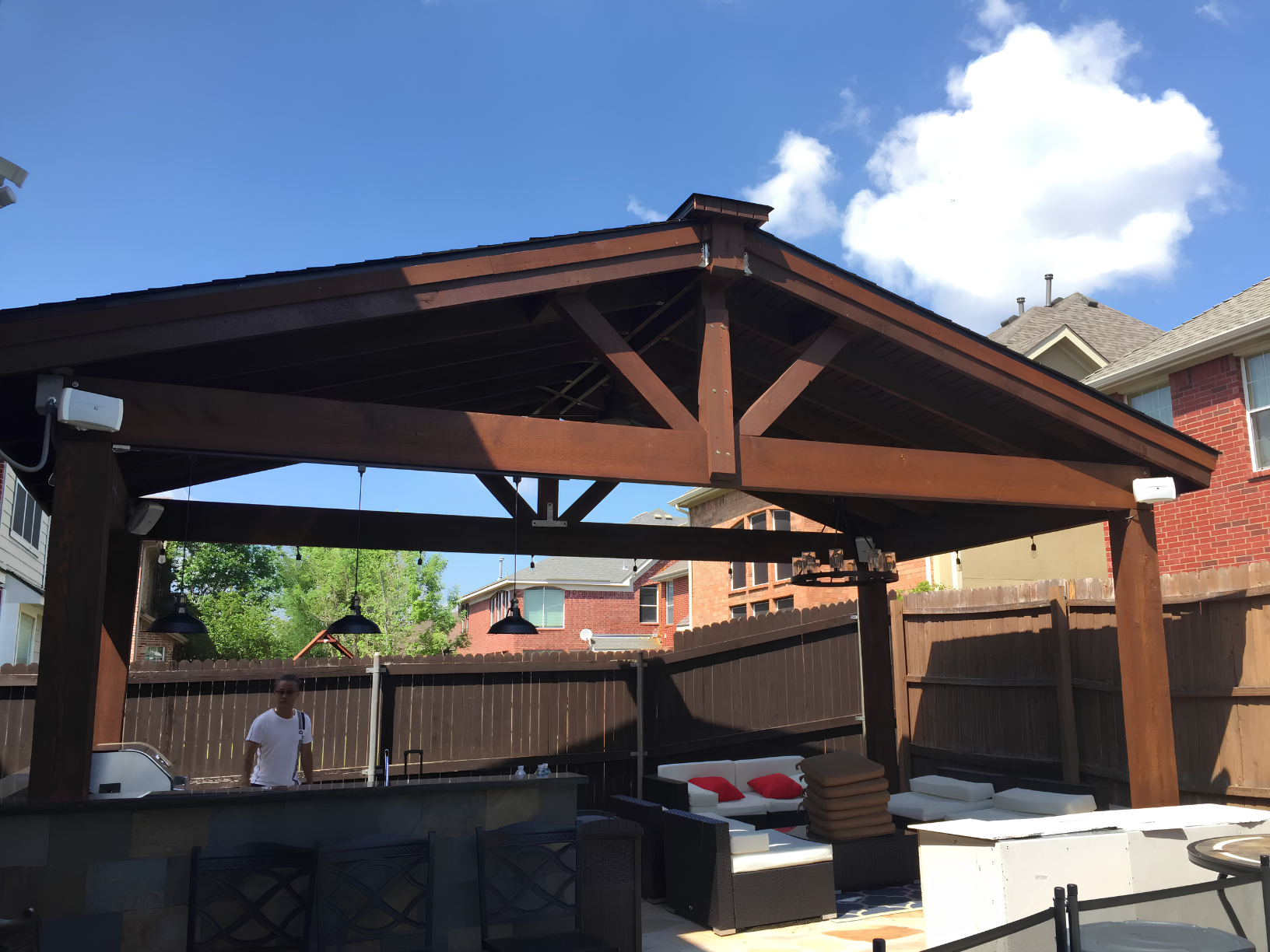 An outdoor patio area with a wooden pergola, string lights, and outdoor furniture including a sectional sofa with red pillows and cushions, under a clear blue sky with some clouds.