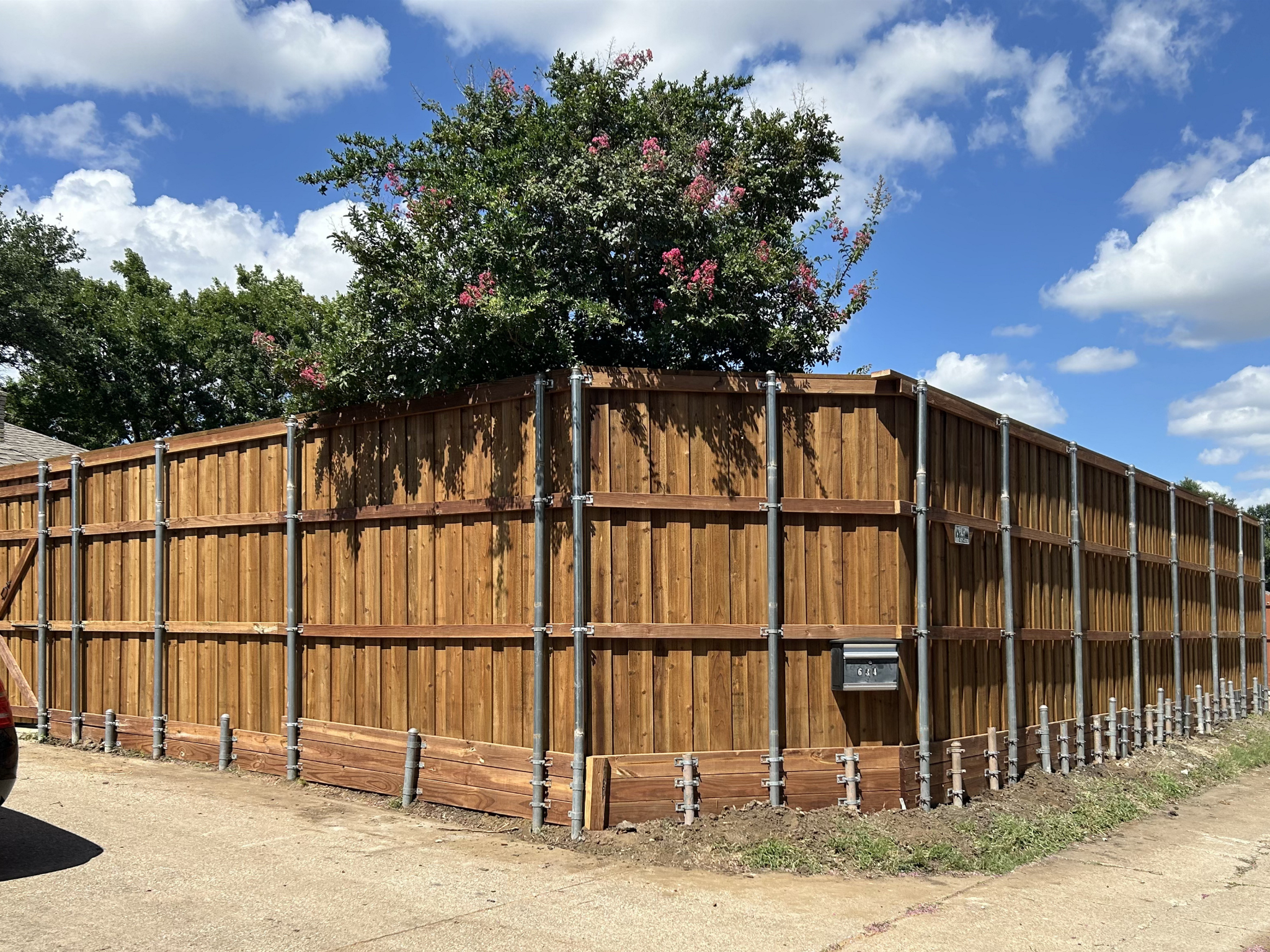 A wooden fence with metal support posts surrounds the yard of a house. There are trees behind the fence, with pink flowers on some of the branches. The sky above is partly cloudy with a bright blue background.