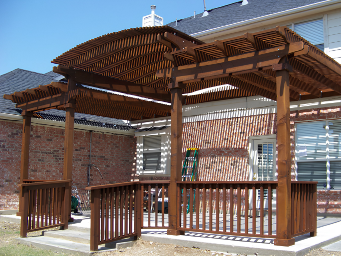 Wooden pergola with slatted roof and railing on a concrete patio in a backyard.
