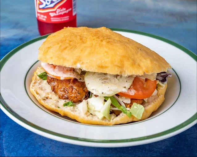 Fried fish sandwich with lettuce, tomato, and tartar sauce on a bun, served on a plate with a soda bottle in the background.