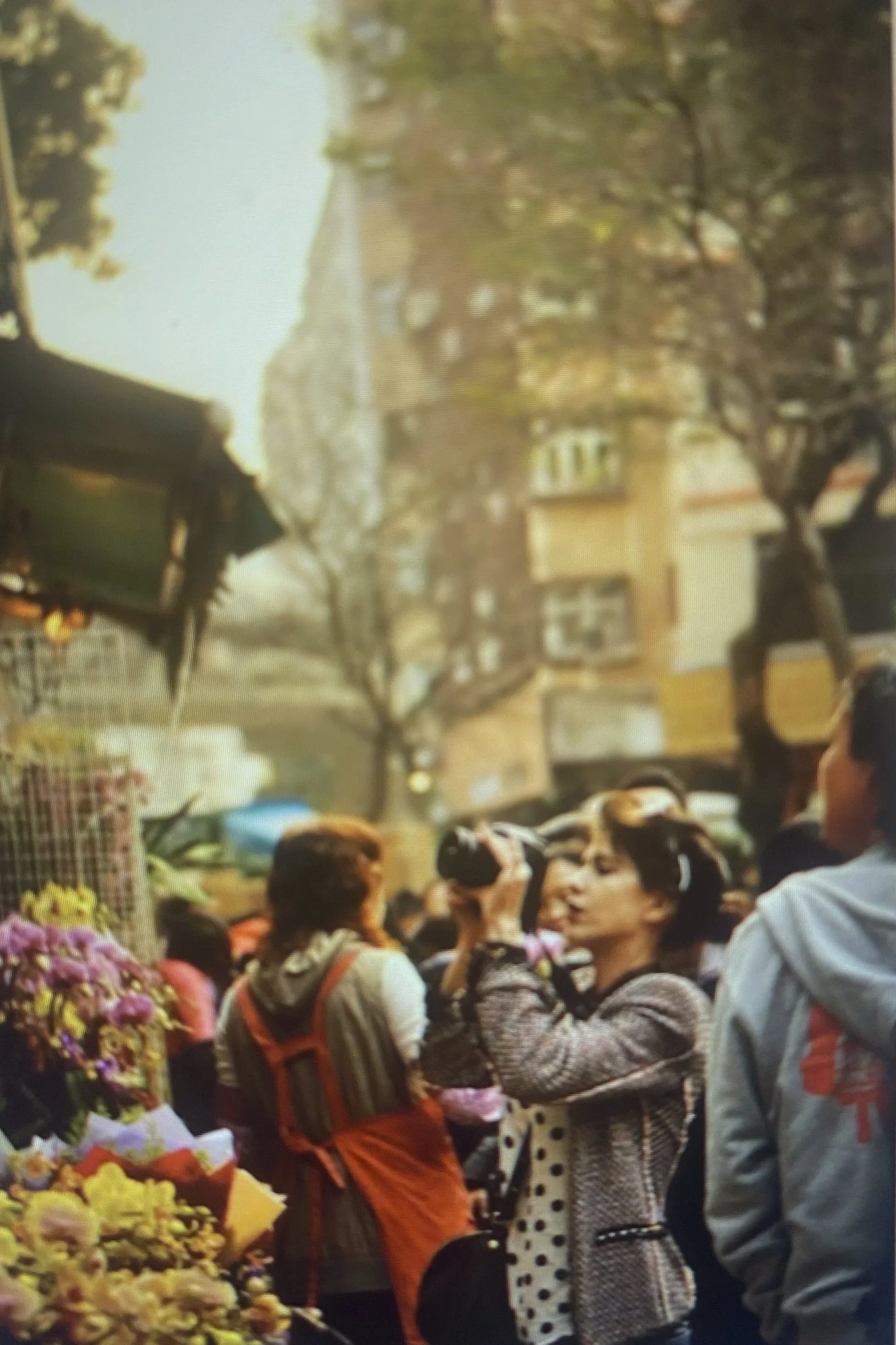 A crowd of people gathered outdoors near a flower stand with colorful flowers, with trees and buildings in the background.