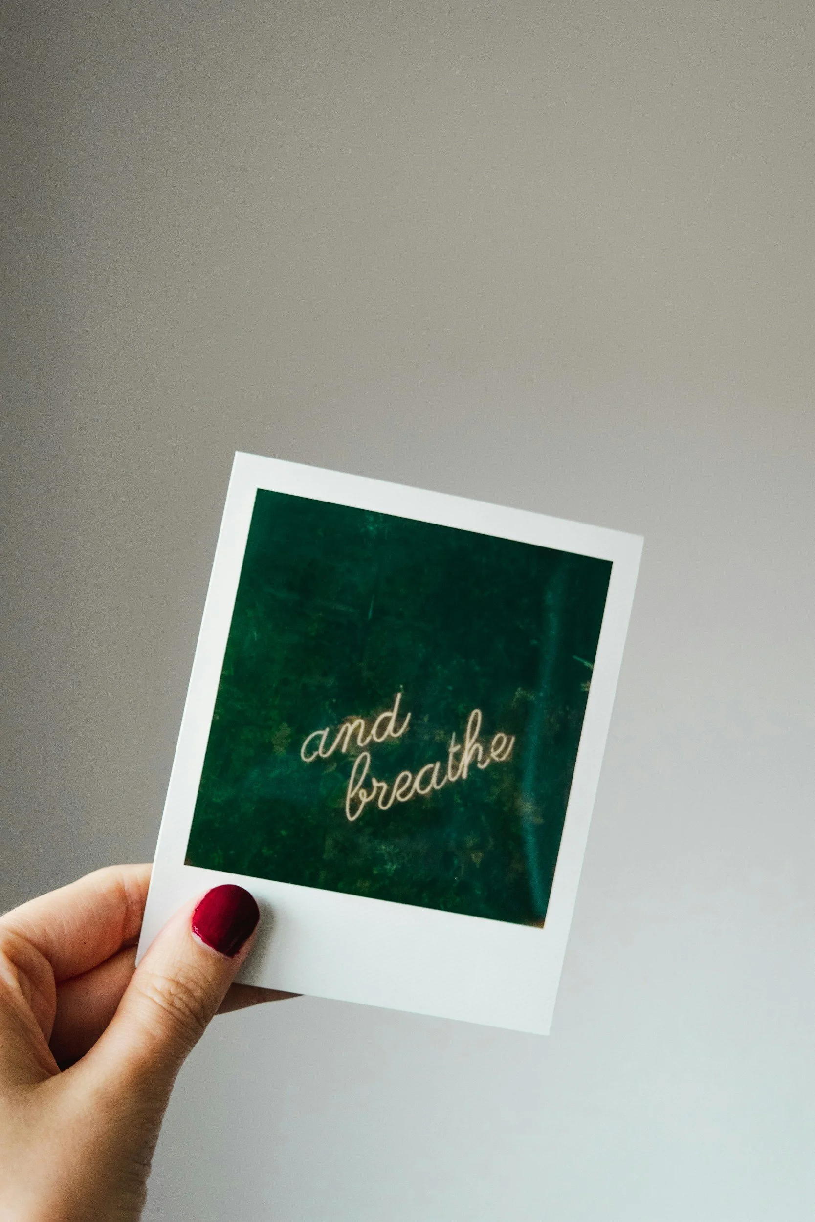 A hand with red painted nails holding a photo with the words "and breathe" written on it, against a plain gray background.