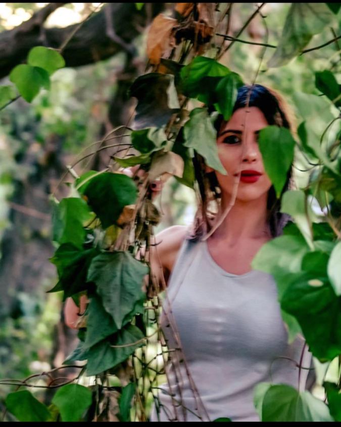 Woman with long dark hair and red lipstick standing among green leafy vines in a forest.