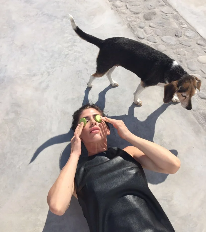 Woman lying on concrete ground with cucumber slices over her eyes, holding her head with both hands, and a dog standing nearby.