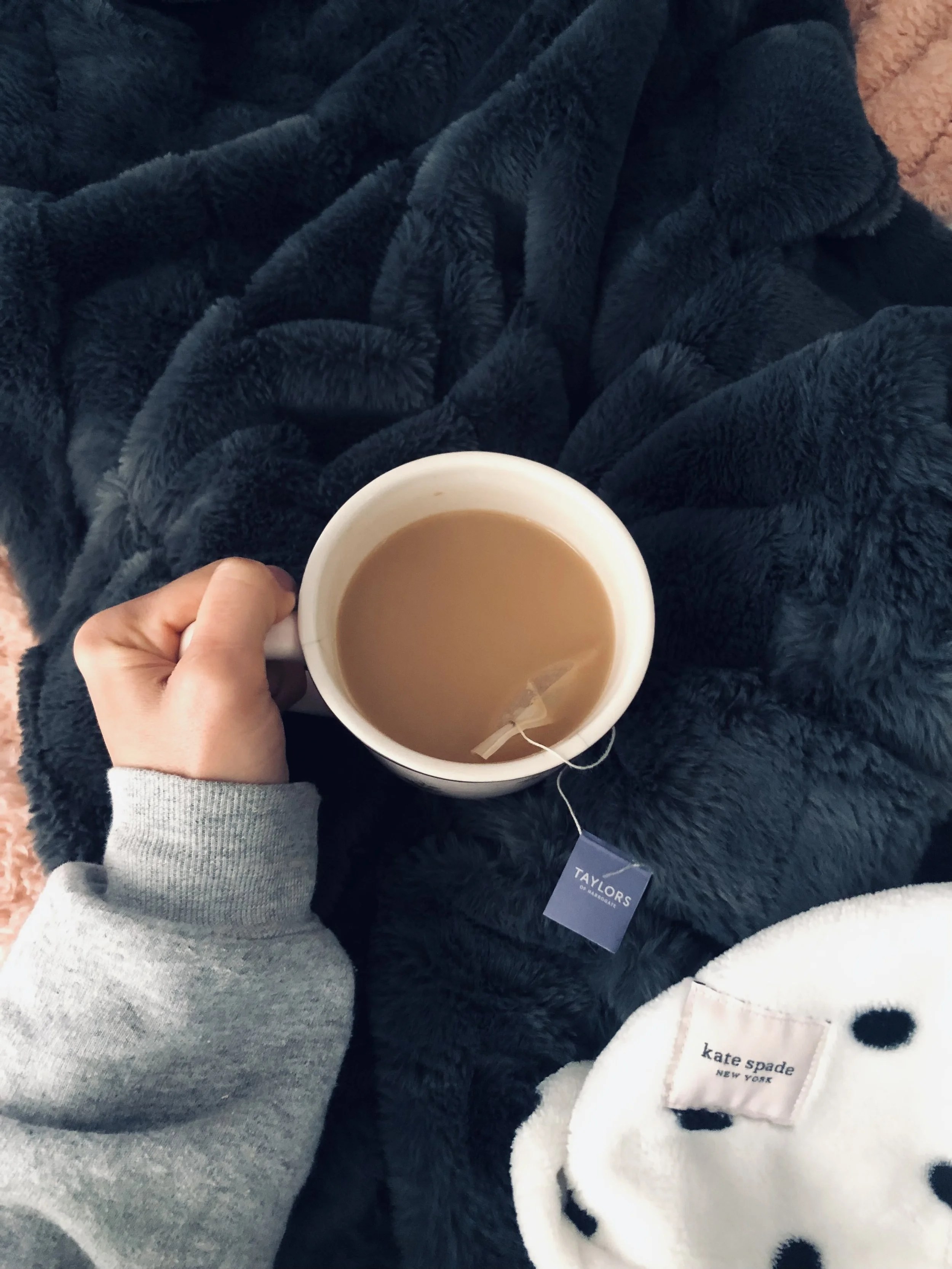 Person holding a white mug with a hot beverage, sitting on a dark fuzzy blanket, wearing a gray sweater and a white Pajama with black polka dots and a Kate Spade tag.