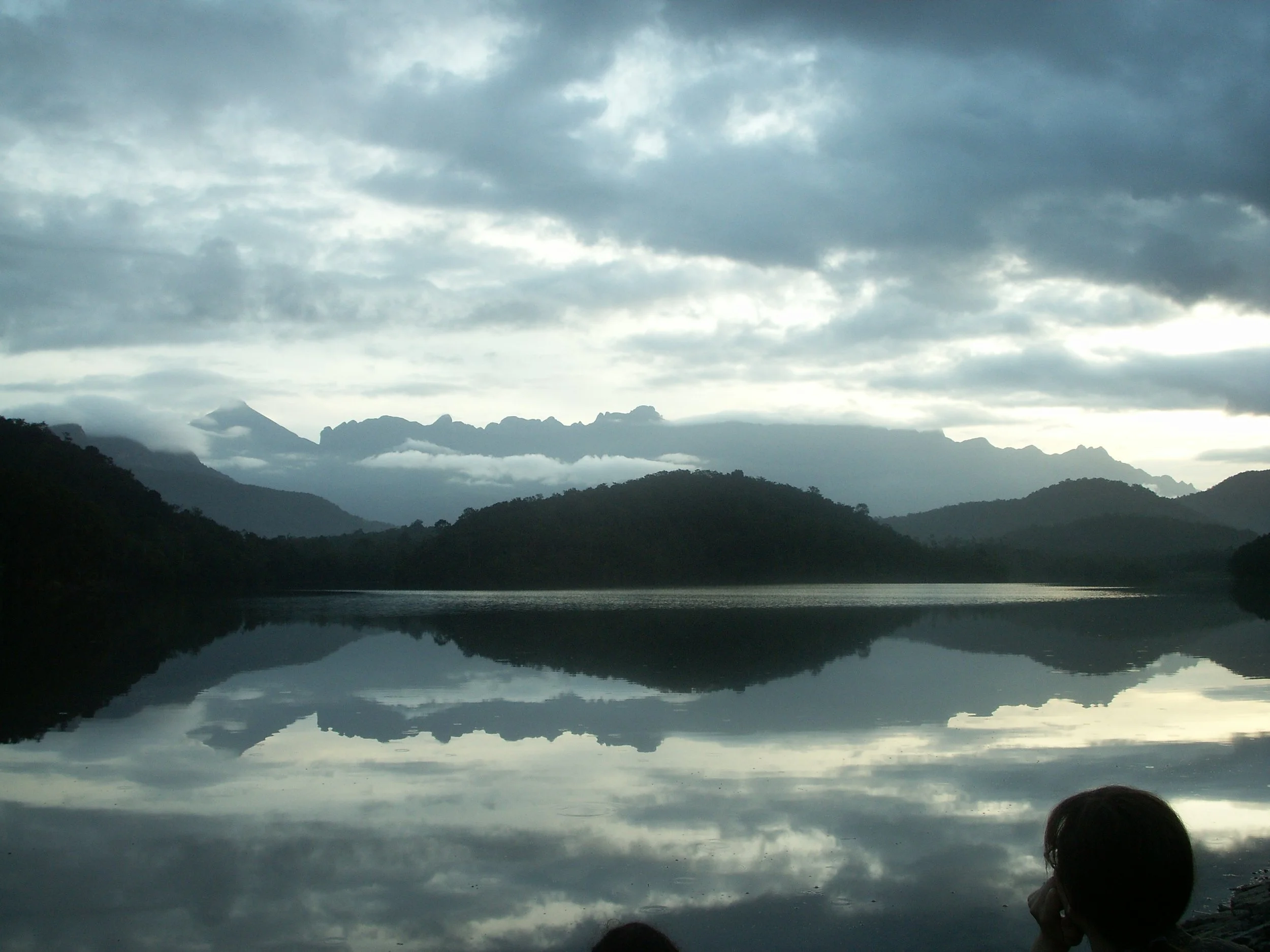 Cloudy sky over mountains and a body of water that reflects the landscape, with some people's heads visible in the foreground.