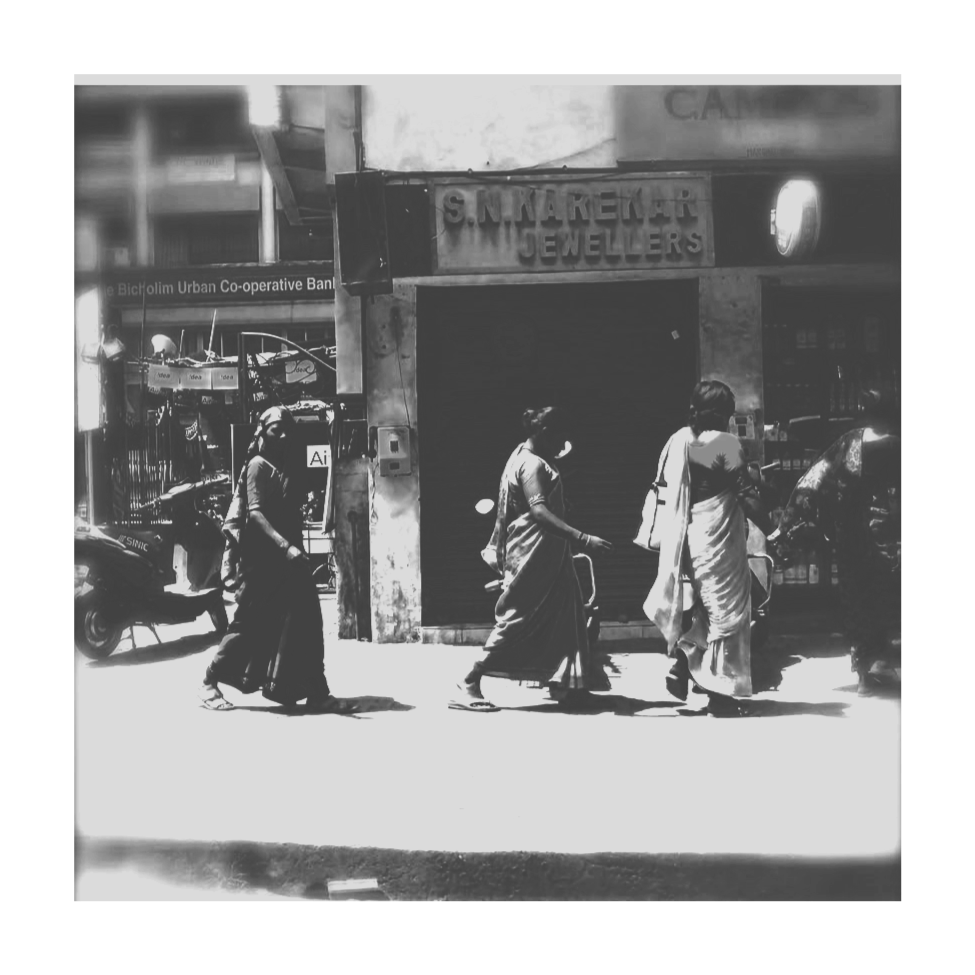 Four women dressed in traditional Indian saris walk on a city street in India, some accompanied by a man, with closed shops and buildings in the background.