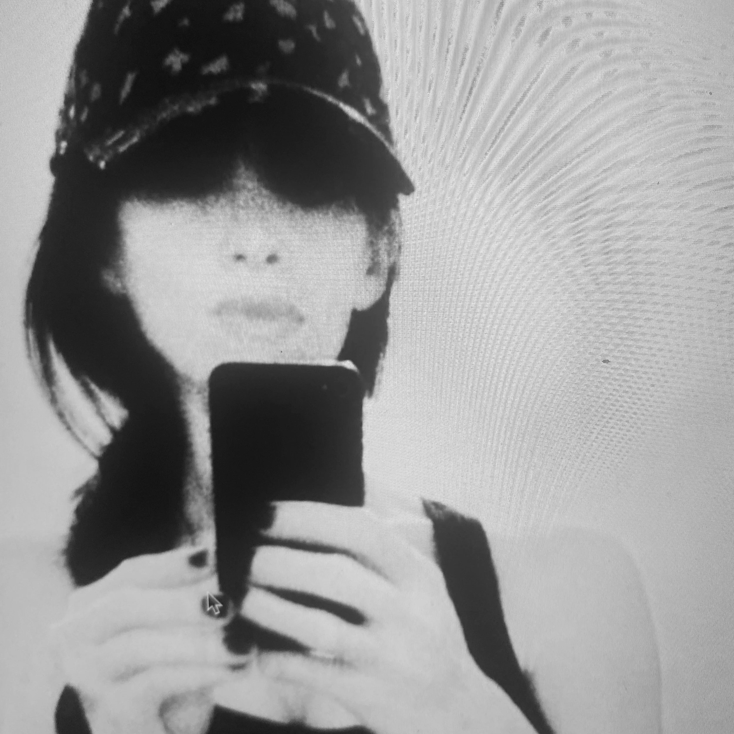 Close-up black-and-white image of a person's face, with dark eye makeup, dark lipstick, short hair, and a distinctive dark clothing pattern. The photo appears to be taken from a side angle with high contrast.