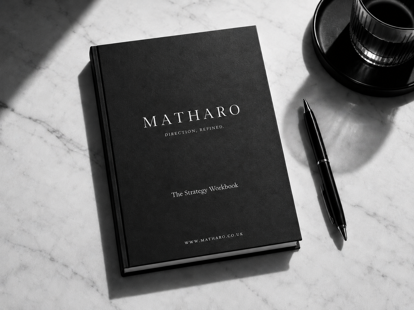 Black strategy workbook titled 'MATHARO: Direction, Refined' on a white marble surface, with a black pen beside it, and a black cup on a black coaster.