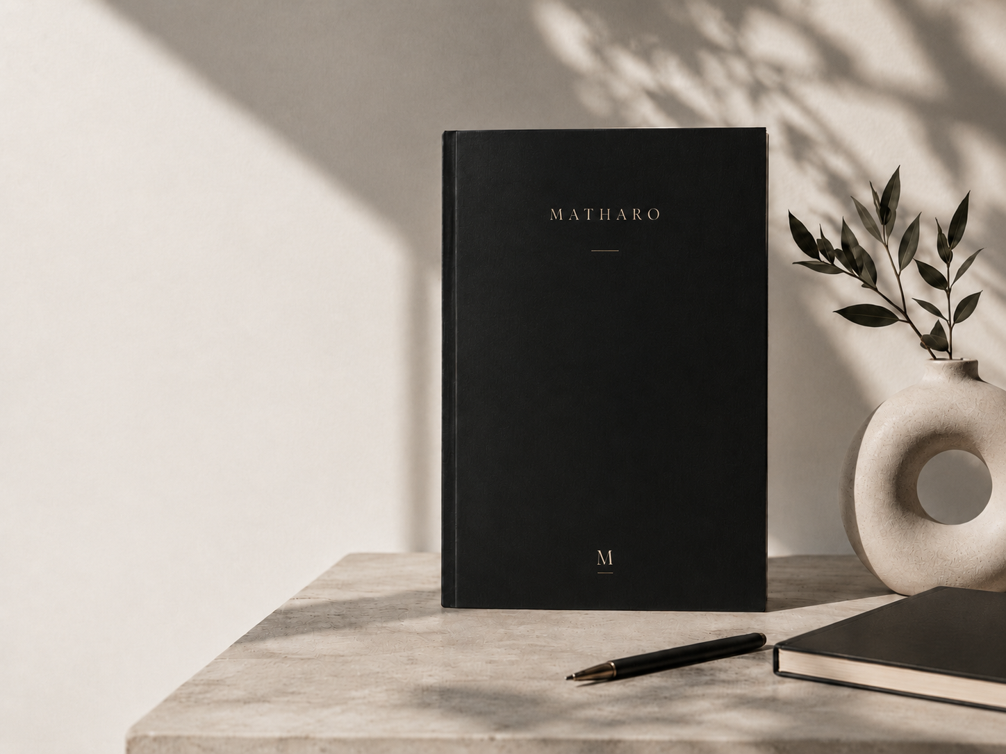 A black hardcover book with the title 'MATHARO' on the cover, a beige ceramic vase with a hole and leafy branch decoration, and a black pen on a light-colored wooden surface, with shadows and sunlight on the wall.