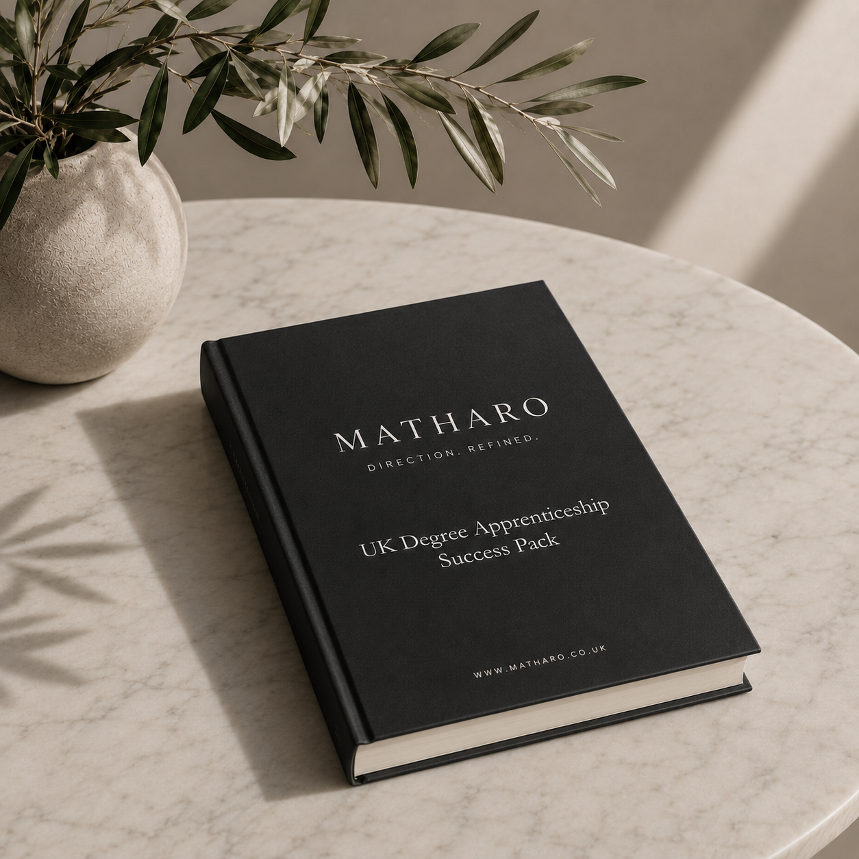 A black book titled "Matharo: Direction. Refined." with subtitle "UK Degree Apprenticeship Success Pack" and website "www.matharo.co.uk" is placed on a light-colored marble table. A beige textured vase with green leafy plant is positioned nearby, and sunlight casts soft shadows on the table.