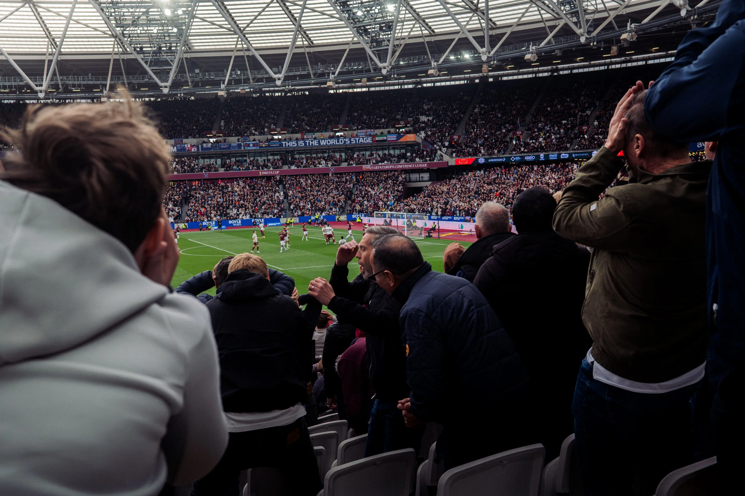 View of a stadium filled with spectators watching a soccer match, with players on the field and a digital banner displaying sponsors and event information.