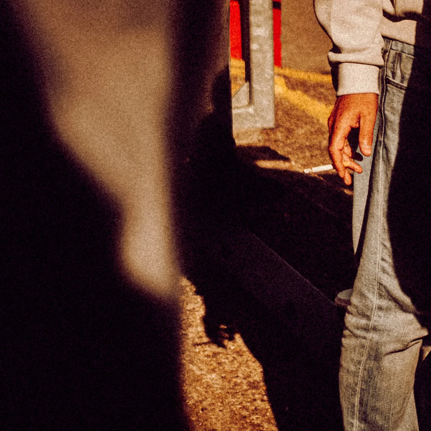 Person wearing gray hoodie and jeans holding a lit cigarette near a sidewalk at a football match.